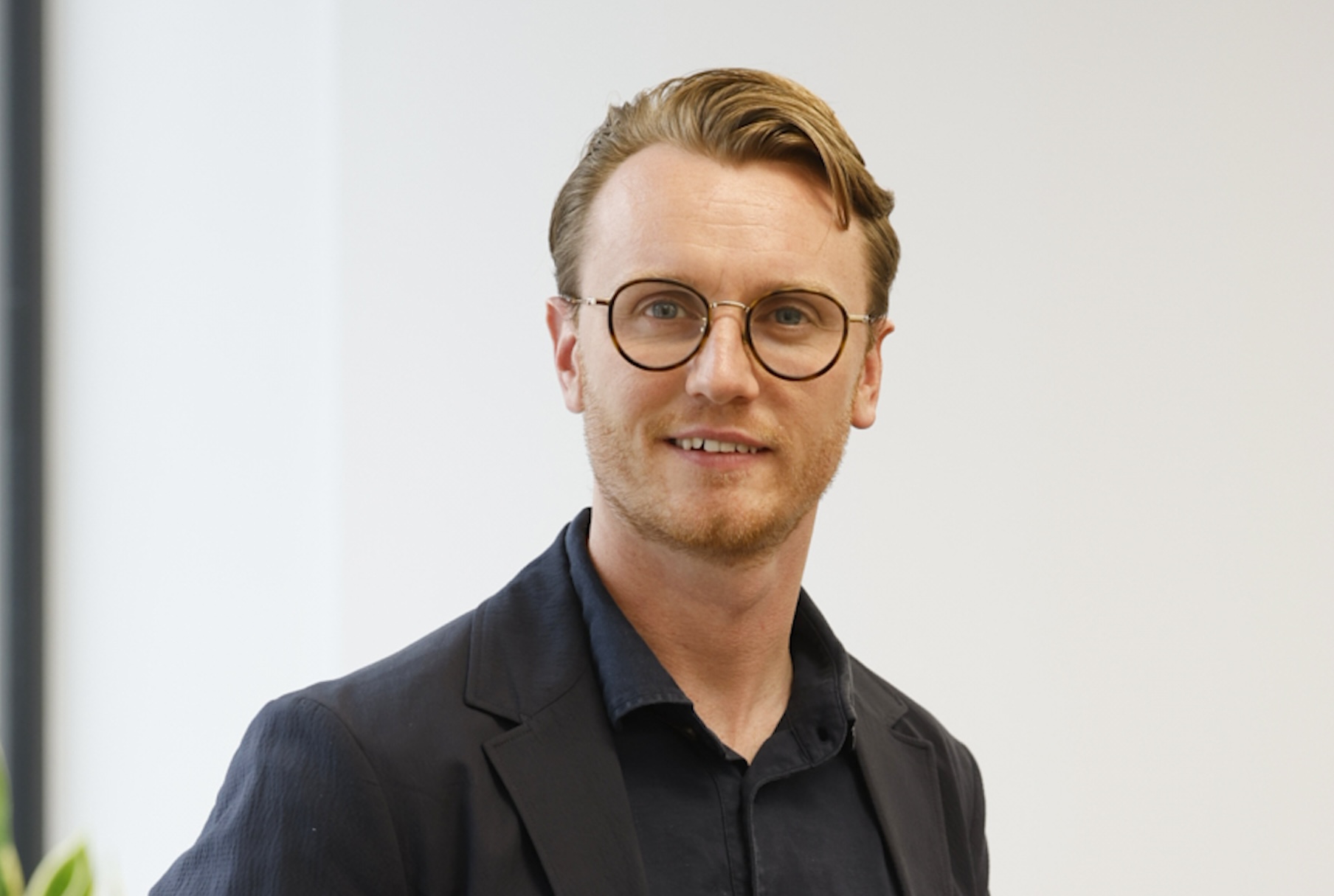 Calum McEwen wears a black shirt, black jacket and glasses. He is smiling at the camera.