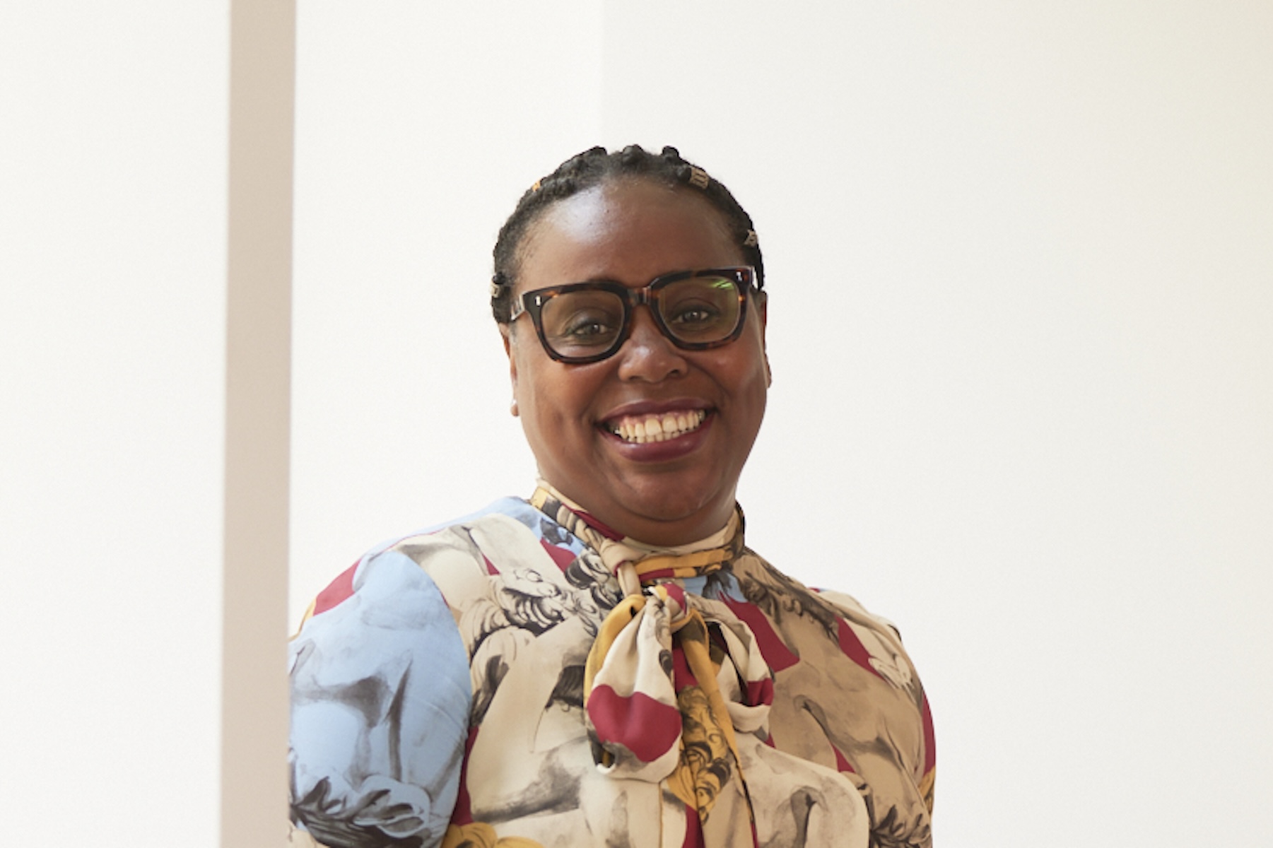 Tracy wears black glasses and a high neck dress that is light beige, dark red and light blue, it ties at the neck with a bow. Tracy is smiling.