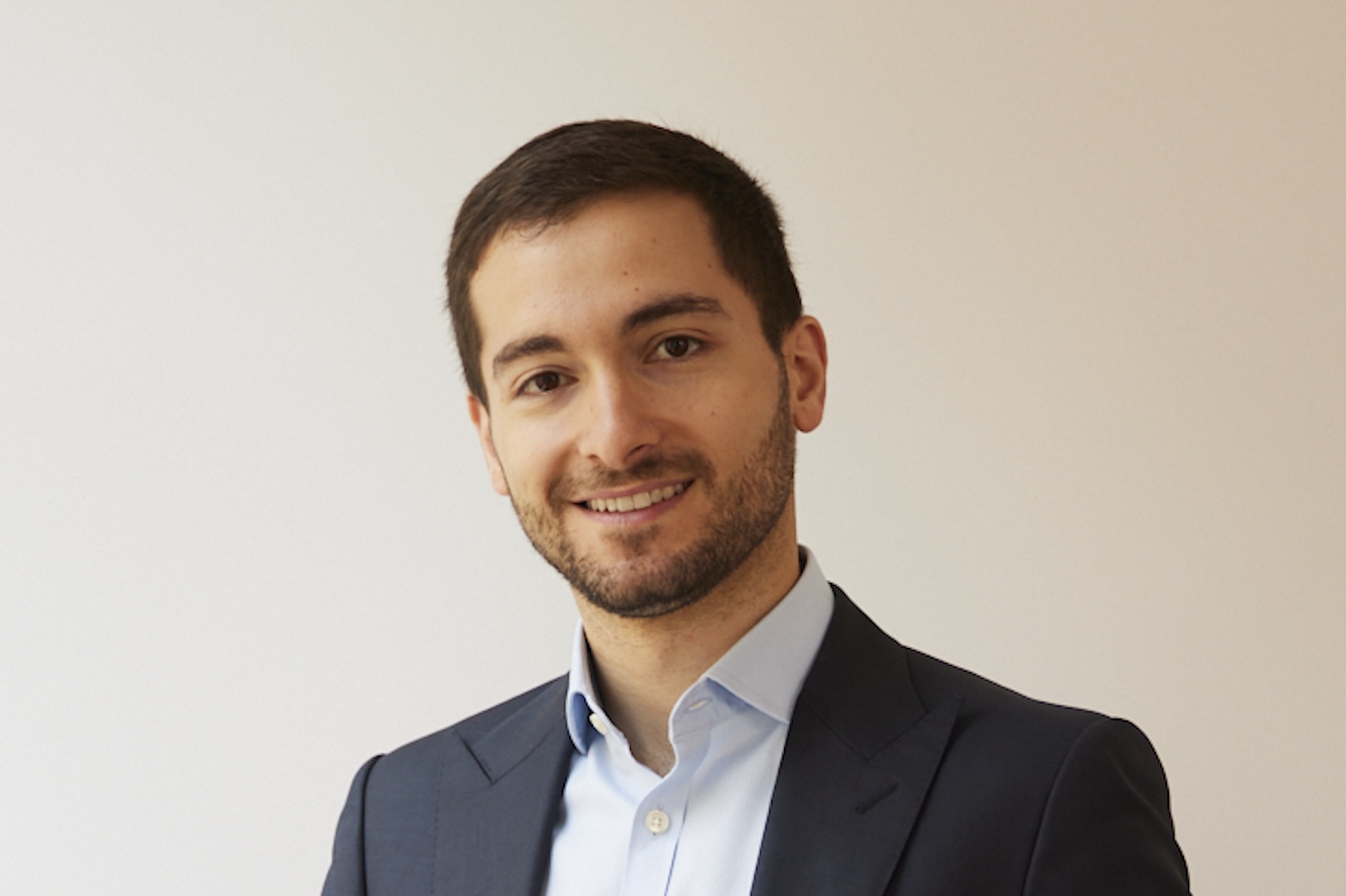 Tigran wears a dark blue jacket and a light blue shirt and is smiling.